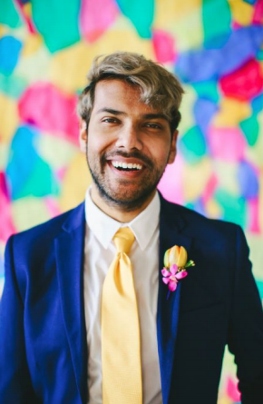 A photo of Tau Zaman smiling, wearing a blue suit with a yellow tie and a boutonnière of yellow and pink flowers. They are standing in front of a vibrant background of yellow, pink, teal, and blue abstract shapes.