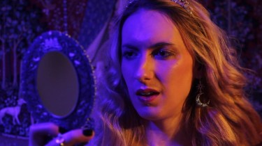 Natalie Wynn, a woman with long blond hair wearing earrings and a tiara, looking into a mirror, lit by deep purple from her right